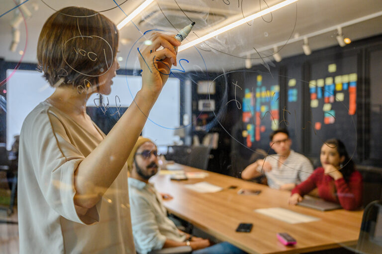 A person drawing diagrams on a glass board with a marker, while colleagues watch from a table in a modern office setting.