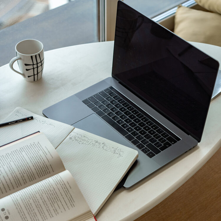 A laptop, a notebook, and an open book on a white table with a striped mug beside them, near a window.