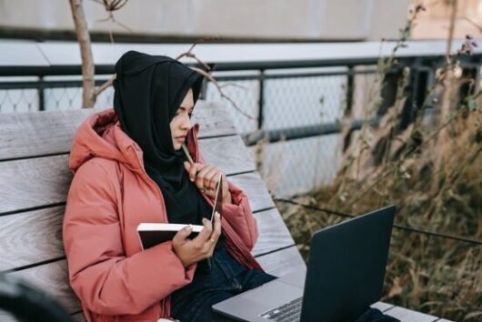 Woman working outside on a bench with her laptop and notebook